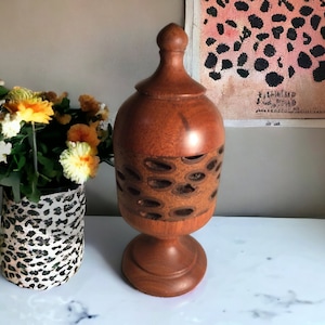 May include: A wooden vase with a carved design and a lid. The vase is brown and has a natural wood finish. The vase is sitting on a white table with a bouquet of yellow flowers in a leopard print vase.