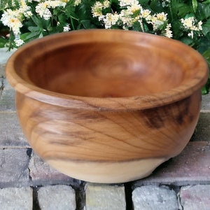 May include: A large, round wooden bowl with a natural wood finish. The bowl is made of a light brown wood with darker brown streaks. The bowl is sitting on a brick surface.