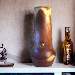 Puede incluir: Un jarrón de madera con un patrón de veta de madera natural. El jarrón es marrón y tiene una forma única. Está sentado en un estante blanco con un pequeño objeto decorativo y una botella de vidrio.