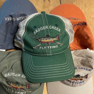 May include: Five different baseball caps with a mesh back. The caps are all green, brown, and tan. The caps have the text "Badger Creek Fly Tying" embroidered on the front.