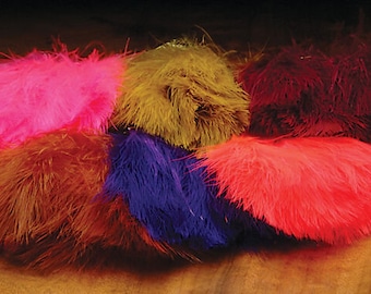 May include: Assortment of ostrich feathers in various colors, including pink, brown, gold, purple, and red. The feathers are arranged in a pile on a wooden surface.