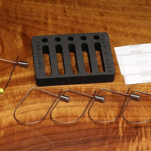 May include: A black plastic beading tool with five slots and the text "FEED A BEAD" on the side. The tool is accompanied by four metal beading loops and three white adhesive strips.
