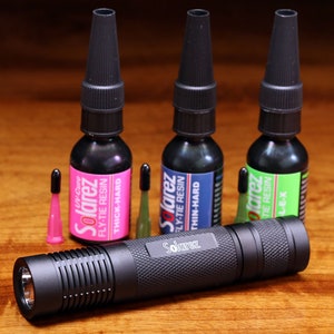 Puede incluir: Tres botellas de resina Solarez para atar moscas, en diferentes colores y consistencias: gruesa y dura, fina y dura, y L-E-X. Una linterna negra con el logotipo de Solarez está en primer plano.