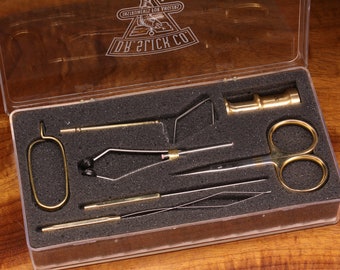 May include: A clear plastic box containing a fly tying kit. The kit includes a bobbin, a pair of scissors, a bodkin, a hackle pliers, and a whip finisher. The box is labeled "D'Brick Co. Implements for Flytiers".