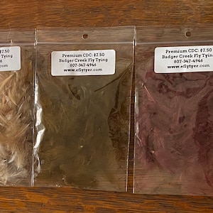 May include: Six bags of CDC feathers in various colors, including white, brown, green, red, and black. The bags are labeled with the price and the name of the company, Badger Creek Fly Tying.