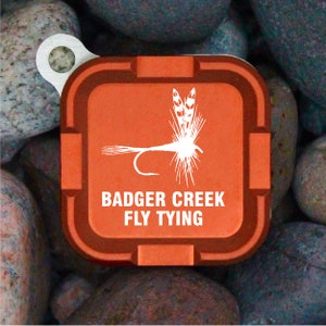 Könnte beinhalten: Eine kleine, quadratische Fliegenbindebox in Orange mit einer weißen Fliegen-Silhouette und dem Text "Badger Creek Fly Tying" auf der Vorderseite.