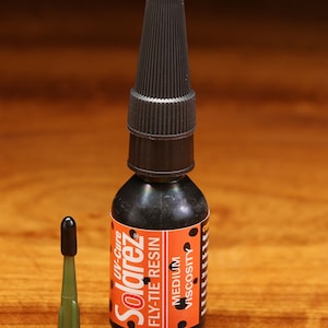 Puede incluir: Una botella negra de resina Solarez UV-Cure Fly-Tie con una tapa negra y una etiqueta blanca con lunares negros. La etiqueta dice "Solarez UV-Cure Fly-Tie Resin" y "Viscosidad media". También se muestra una botella verde más pequeña con una tapa negra.