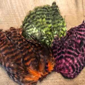 May include: Three bundles of feathers in shades of brown, green, and purple. The feathers have a striped pattern and are arranged on a light brown surface. The feathers are likely for crafting or fly tying.