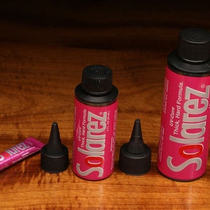Puede incluir: Tres botellas de resina Solarez UV-Cure Thick, Hard Formula, dos en botellas de 2 fl oz y una en un tubo más pequeño. Las botellas son rosas con letras plateadas y tapas negras. El tubo es rosa con letras plateadas y una tapa negra.