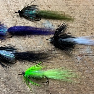 May include: Four different fly fishing lures with various colored feathers and tails. The lures are arranged in a row on a wooden surface.