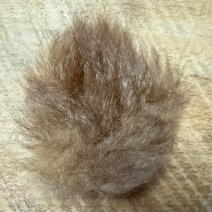May include: A close-up shot of a small, round, light brown fur ball. The fur appears soft and fluffy, with varying shades of brown and tan. The texture is dense and slightly irregular, suggesting a natural material.