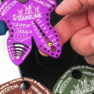 Pode incluir: Ferramenta de amarra&ccedil;&atilde;o de moscas Hareline Keough roxa com uma variedade de medidas para tamanhos de anzol, tamanhos de contas e tamanhos de penas. A ferramenta &eacute; feita de metal e tem um logotipo de coelho. O texto "Hareline" e "Keough" est&aacute; impresso na ferramenta.