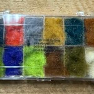 Puede incluir: Un kit transparente para atar moscas con una variedad de fibras sintéticas coloridas. El kit contiene una gama de colores, incluyendo verde, azul, rojo y marrón. El texto "Ranger Creek Fly Tying" es visible en el kit.