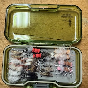 May include: A green fly fishing box with a grey foam insert containing a variety of colourful flies. The box has the text "SFA Laing Budden Creek" printed on the lid.