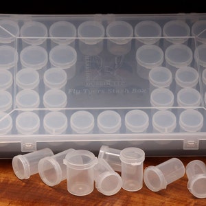 May include: A clear plastic box with 36 small clear plastic containers, labeled "Fly Tyers Stash Box". The box is open and some of the containers are scattered on a wooden surface.