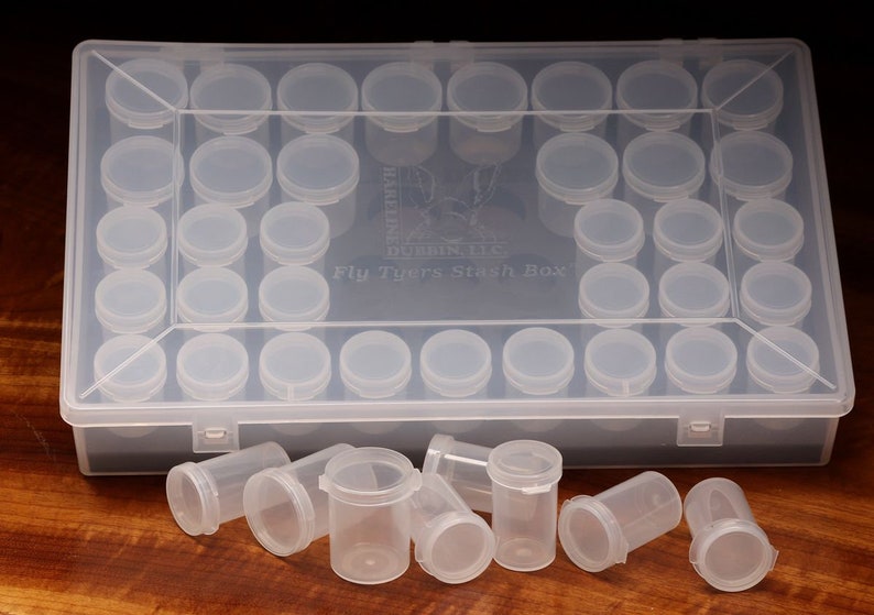 May include: A clear plastic box with 36 small clear plastic containers, labeled "Fly Tyers Stash Box". The box is open and some of the containers are scattered on a wooden surface.