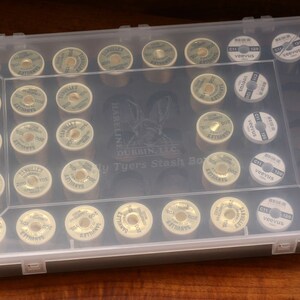 May include: A clear plastic box filled with spools of thread. The spools are labeled "Hareline Dubbin, LLC" and "Tyers Stash Box".