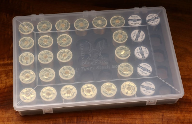May include: A clear plastic box filled with spools of thread. The spools are labeled "Hareline Dubbin, LLC" and "Tyers Stash Box".