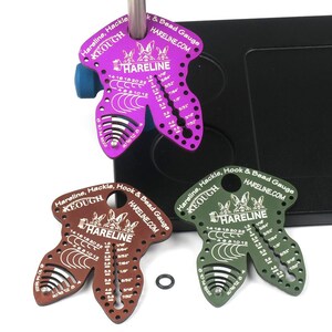 Pode incluir: Tr&ecirc;s ferramentas de amarra&ccedil;&atilde;o de moscas de metal de diferentes cores, cada uma com a forma de um coelho com o texto "Hareline, Hackle, Hook & Bead Gauge" e "HARELINE.COM". As ferramentas t&ecirc;m marcas para medir os tamanhos dos ganchos, os tamanhos das contas e os tamanhos das penas.