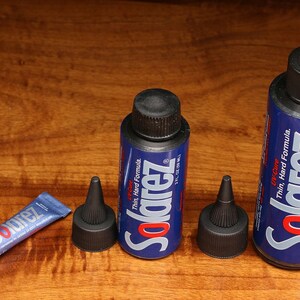Puede incluir: Tres botellas de resina UV Solarez para proteger señuelos de pesca. Las botellas son azules y negras con texto blanco. El texto en las botellas dice "Solarez", "Fórmula fina y dura", y "UV Cure". Las botellas están sobre una superficie de madera.