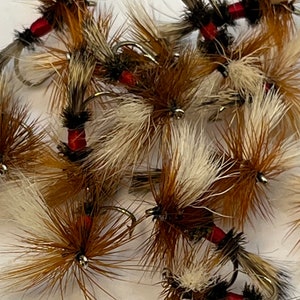 May include: A close-up of a group of brown and white fly fishing lures with red accents. The lures are tied with feathers and fur.