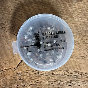 May include: A clear plastic container filled with a variety of fly tying materials. The container has a black label with the text "Badger Creek Fly Tying" and contact information for the business.