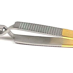 May include: A pair of tweezers with a gold and silver finish. The tips are black and hold a small, gold-colored object. The handle has a textured grip for better control. The tweezers are designed for precision work.