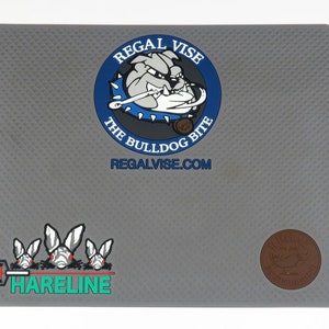 May include: Gray rubber fly tying mat with a bulldog logo that says "Regal Vise The Bulldog Bite Regalvise.com". The mat also has a Hareline logo and a small brown leather patch with the word "Regal" on it.