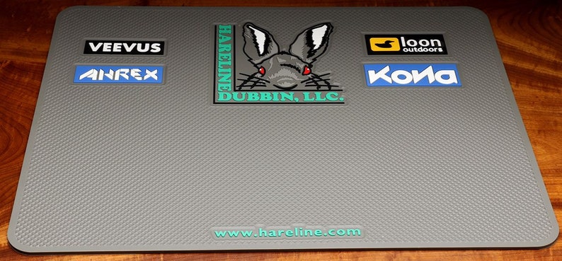 May include: Gray textured rubber mat with a black rabbit logo, the text "Hareline Dubbin, LLC." and the website "www.hareline.com". The mat also has several other logos including "Veevus", "Anrex", "loon outdoors", and "Kona".