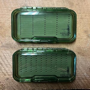 Op de afbeelding: Twee groene plastic vliegenbinddozen met doorzichtige deksels. De dozen hebben een rasterpatroon aan de binnenkant en de tekst "Badger Creek Fly Tying" aan de voorkant.