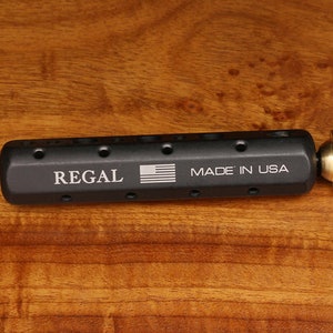 May include: A black metal tool with the words "REGAL" and "MADE IN USA" engraved on it. The tool has a round, gold-colored knob on one end.