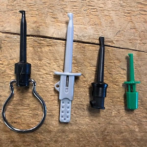Puede incluir: Cuatro sondas de prueba eléctricas de diferentes colores: negro, gris y verde. Una sonda negra tiene un lazo de metal plateado. Se utilizan para pruebas y mediciones eléctricas.