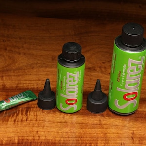 Puede incluir: Tres botellas de Solarez UV-Cure Flex Formula en diferentes tamaños. Las botellas son verdes con tapas y etiquetas negras. La botella más pequeña es un tubo con una tapa verde y negra.