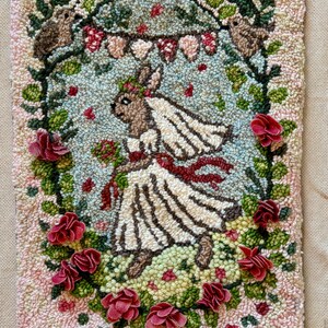 May include: A handmade rug featuring a rabbit bride in a white dress, veil, and holding a bouquet. The design is surrounded by a floral arch with red roses and two rabbits. The background is a mix of light blue, green, and pink.