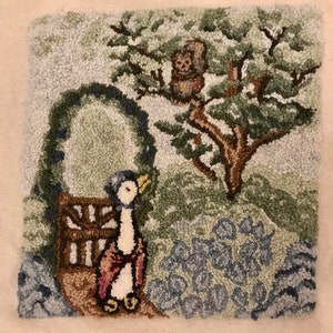 May include: A hand-stitched textile art piece depicting a whimsical garden scene. A penguin-like figure stands near a wooden gate, with a tree and an owl perched in its branches. The piece uses shades of green, blue, brown, and white.