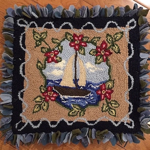 May include: Square, handmade rug featuring a sailboat on the water, surrounded by red flowers and green leaves. The rug has a beige background, a dark blue border, and a fringe of blue and green fabric around the edges.