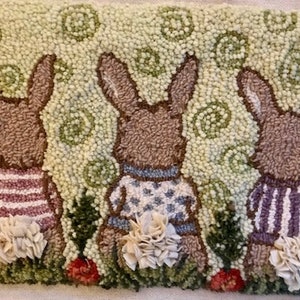 May include: Hand-tufted rectangular rug featuring three brown bunnies wearing sweaters. The bunnies have fluffy white tails and are set against a light green background with floral accents. The bunnies' sweaters are striped and patterned.