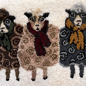 May include: A decorative rug depicting three sheep with textured wool in brown, tan, and grey. Each sheep wears a scarf in red, brown, and gold. The background is a neutral off-white. A charming, handcrafted item.