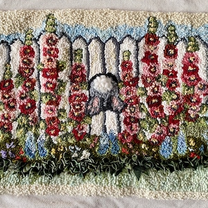 May include: Hand-tufted rug featuring a white picket fence with vibrant red and pink hollyhocks. A fluffy white bunny tail peeks through the fence. The rug has a light blue border and a textured, off-white background, creating a charming garden scene.