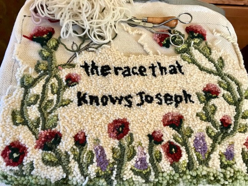 K&ouml;nnte beinhalten: Ein handgefertigter Teppich mit dem Text "the race that knows Joseph" in Schwarz. Der Teppich hat einen floralen Rand mit roten und lila Blumen und gr&uuml;nen Bl&auml;ttern. Eine Schere und Garn liegen obenauf.