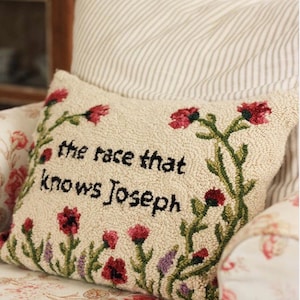 K&ouml;nnte beinhalten: Cremefarbenes, strukturiertes Kissen mit gesticktem Text "the race that knows Joseph" und floralen Akzenten. Die Blumen sind rot und lila mit gr&uuml;nen Stielen. Das Kissen liegt auf einem floral gemusterten Stuhl.