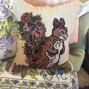 May include: A decorative square pillow with a textured, off-white background. The pillow features a detailed image of a brown squirrel holding a nut, with a floral accent of purple and green fabric. The pillow is approximately 16 inches square.