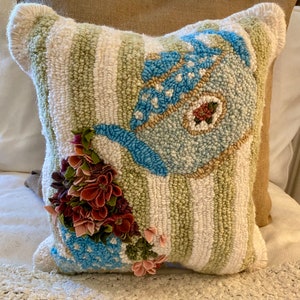 May include: A decorative pillow with a hand-tufted design. The pillow features a bird motif in shades of blue, beige, and brown, set against a backdrop of vertical green and white stripes. A cluster of fabric flowers in burgundy and pink adds a 3D element.