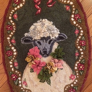 May include: Oval-shaped rug featuring a sheep design. The sheep has a fluffy, light gray head and is adorned with a floral garland. The rug's border is decorated with flowers and a red ribbon, set against a dark green background.