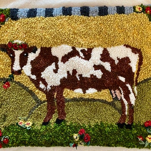 Peut inclure: Tapis crocheté à la main représentant une vache marron et blanche avec une couronne de fleurs, dans un champ vert. Le fond est jaune, avec une bordure de fleurs et un motif à carreaux noirs et gris. Un charmant élément de décoration rustique.