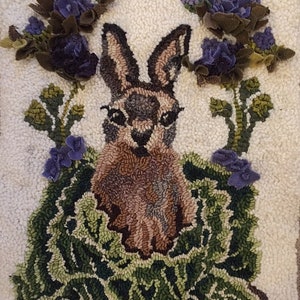 May include: A handcrafted rug featuring a rabbit surrounded by green foliage and purple flowers. The rabbit has brown and tan fur, with large ears. The flowers and foliage are made of textured fabric, set against a white background.