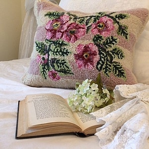 May include: A square cushion with a floral design of pink roses and green leaves. The cushion is on a white bed with an open book and white lace.