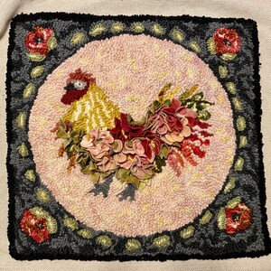May include: A square, handmade rug featuring a rooster design. The rooster is in the center, surrounded by a pink circle and a dark gray border with floral accents. The rooster has a yellow body, red comb, and is adorned with fabric flowers.