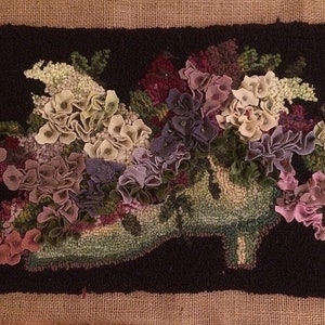 May include: A handmade rug featuring a floral design. The rug depicts a high-heeled shoe overflowing with colorful flowers in shades of pink, purple, green, and white against a black background. The rug is rectangular and has a textured appearance.