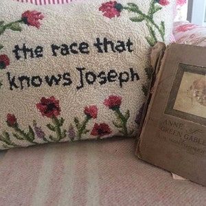 K&ouml;nnte beinhalten: Ein dekoratives Kissen mit dem Text "the race that knows Joseph", umgeben von floralen Stickereien. Daneben liegt ein Vintage-Buch mit dem Titel "Anne of Green Gables" von L.M. Montgomery mit verblichenem Einband.